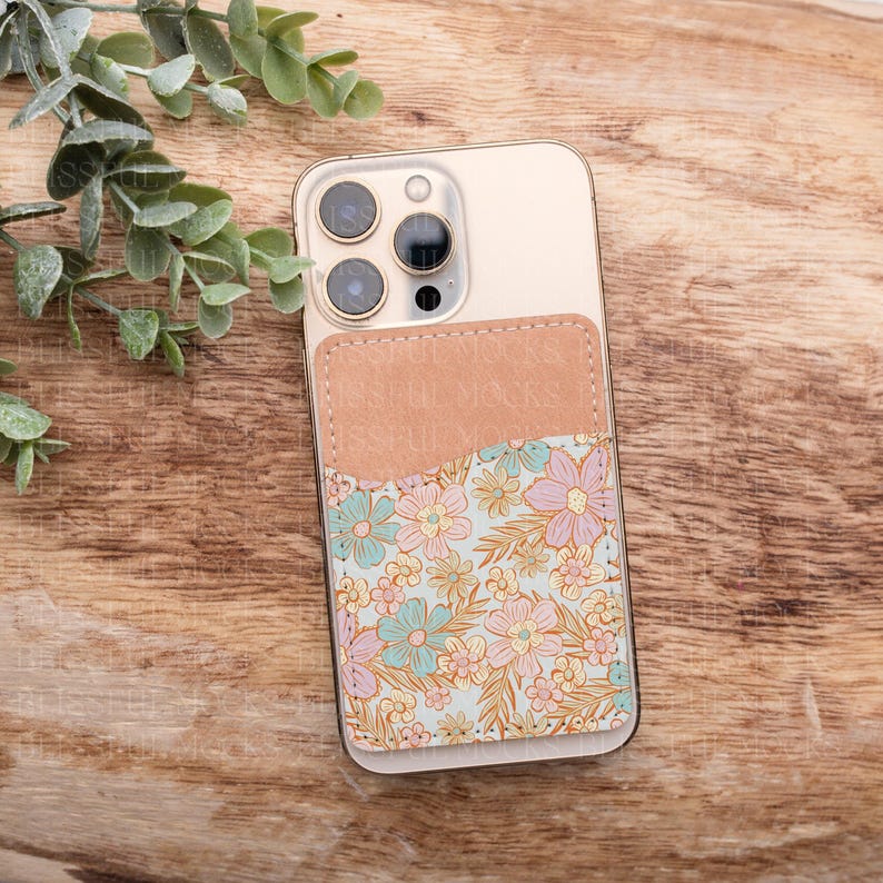 May include: A white phone case with a brown leather pocket on the back. The pocket has a floral pattern with pink, blue, and yellow flowers.