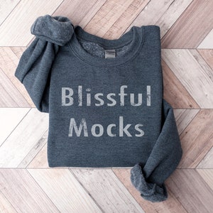Pu&ograve; includere: Una felpa grigia scura con la scritta "Blissful Mocks" stampata in bianco sul davanti.