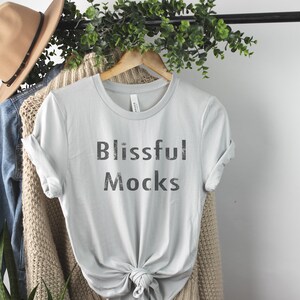 K&ouml;nnte beinhalten: Ein hellgraues T-Shirt mit dem Aufdruck "Blissful Mocks" auf der Vorderseite. Das Shirt h&auml;ngt an einem Holzb&uuml;gel und ist von Gr&uuml;npflanzen umgeben.