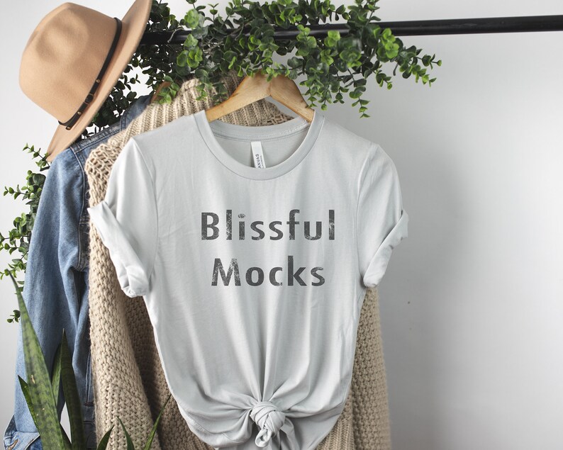 K&ouml;nnte beinhalten: Ein hellgraues T-Shirt mit dem Aufdruck "Blissful Mocks" auf der Vorderseite. Das Shirt h&auml;ngt an einem Holzb&uuml;gel und ist von Gr&uuml;npflanzen umgeben.