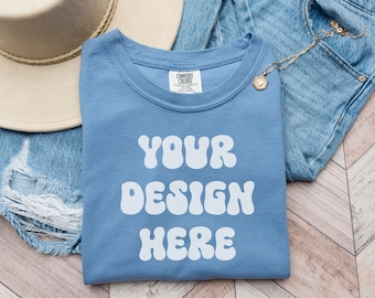 May include: A blue t-shirt with the text "YOUR DESIGN HERE" in white, retro-style lettering. The shirt is folded and laid on a wooden surface with a pair of blue jeans and a brown hat.