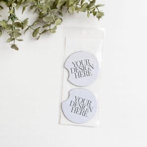May include: Two white round coasters with a cut-out for a cup. The coasters have the text "YOUR DESIGN HERE" printed on them.