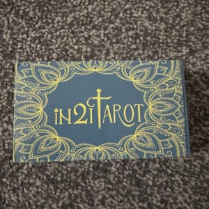 Op de afbeelding: Een rechthoekige tarotkaartdoos met een blauwe achtergrond en een gouden bloemenrand. De tekst "IN 2 TAROT" staat in gouden letters. De doos ligt op een grijs, getextureerd oppervlak.