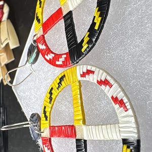 May include: A pair of colorful earrings featuring a circular design with segments of red, yellow, black, and white. The earrings have a woven texture and are suspended from silver-tone ear wires. The design is reminiscent of a Native American medicine wheel.