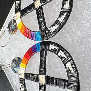 May include: A pair of handmade earrings featuring a circular design with a cross in the center. The earrings have a black and white pattern, with a rainbow gradient on the outer edge. The earrings are suspended from silver-colored hooks.