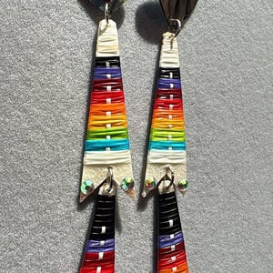 May include: A pair of dangle earrings with heart-shaped tops and colorful, layered, triangular pendants. The pendants feature a rainbow of colors, including red, orange, yellow, green, blue, and purple, with white and black accents. Small, iridescent gems adorn the bottom.
