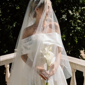 Pode incluir: Uma noiva com um vestido de noiva branco com os ombros descobertos, segurando um buquê de callas brancas. Um véu transparente cobre o rosto da noiva. O fundo apresenta uma varanda de pedra e vegetação exuberante.