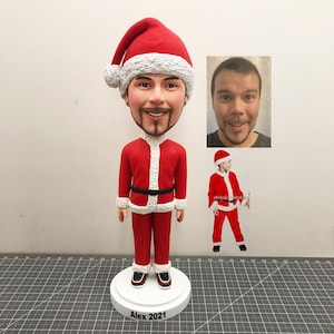 May include: A custom-made Santa Claus figurine with a red suit, white trim, and a black belt. The figurine has a large head with a realistic face and a Santa hat. The figurine is standing on a white base with the name "Alex 2021" printed on it.