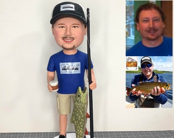 May include: A custom bobblehead figurine of a person wearing a blue shirt, khaki shorts, and a black baseball cap. The person is holding a fishing rod and a fish. The bobblehead is on a white base.