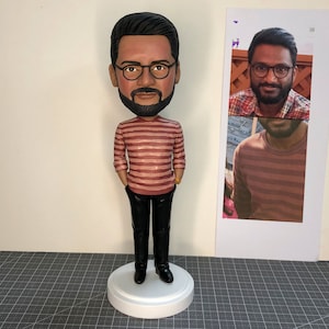 May include: A custom bobblehead figurine of a person with a beard wearing glasses and a red and white striped shirt with black pants. The figurine is standing on a white base.