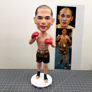 Könnte beinhalten: Eine personalisierte Bobblehead-Figur eines Mannes, der rote Boxhandschuhe und schwarze Shorts mit dem Aufdruck "Taipan" trägt. Die Figur steht auf einem weißen Sockel mit dem Aufdruck "Dumpling Forever". Die Figur ist in einer Boxhaltung dargestellt.
