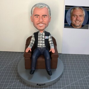 May include: A custom figurine of a person sitting in a brown armchair. The person is wearing a black vest over a plaid shirt, blue jeans, and black shoes. The figurine is on a gray base with the name "Agari" written on it. The person is wearing a badge that says "Blood Drive Volunteer".