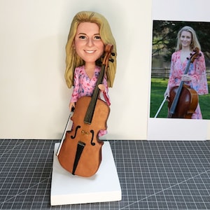 May include: A custom-made figurine of a person holding a cello. The figurine is wearing a pink and white floral dress. The cello is brown with a black bow.