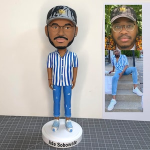 Könnte beinhalten: Ein blau-weiß gestreiftes Hemd, blaue Jeans und eine graue und schwarze Camouflage-Baseballkappe. Die Figur steht auf einem weißen Sockel mit dem Namen "Ade Sobowale" darauf.