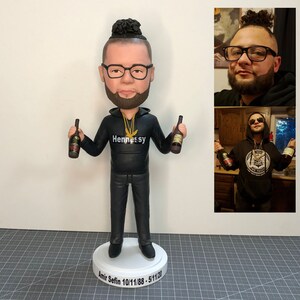 May include: A custom-made figurine of a person with a beard and a bun in their hair. The figurine is wearing a black hoodie with the word "Hennessy" on it and holding two bottles of Hennessy. The figurine is standing on a white base with the name "Amir Sefin" and the dates "10/11/88 - 5/11/20" written on it.