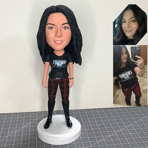 Könnte beinhalten: Eine maßgefertigte Figur einer Person mit langen schwarzen Haaren, die ein schwarzes T-Shirt mit dem Aufdruck "TRUMP 2020", eine rot-schwarze Karohosen und schwarze Stiefel trägt. Die Figur steht auf einem weißen Sockel.