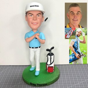 Könnte beinhalten: Eine maßgefertigte Golf-Bobblehead-Figur eines Menschen, der ein blaues Hemd, eine weiße Hose und einen weißen Hut mit dem Text "Retired 2/8/2021" trägt. Die Figur hält einen Golfschläger und hat eine Golftasche mit Schlägern an ihrer Seite. Der Sockel der Figur ist grün und trägt den Namen "Greg Thorson".