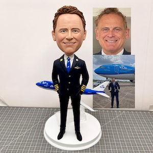 May include: A custom bobblehead figurine of a pilot wearing a blue uniform with gold accents and a blue tie. The figurine is holding a model airplane with the KLM logo and the words "KLM" on the side. The figurine is standing on a white base.