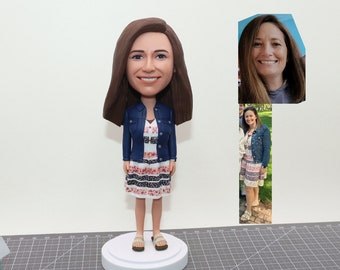 May include: A custom bobblehead figurine of a person with brown hair wearing a blue denim jacket over a white and pink striped dress. The figurine is standing on a white base.