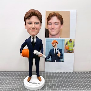 May include: A figurine of a person in a navy blue suit, black tie, and brown shoes, holding an orange hard hat and a rolled-up blueprint. The figurine has a large head and is standing on a white base with text. There are three photos in the background.