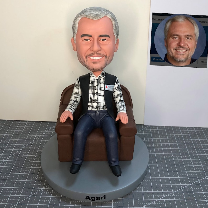 May include: A custom figurine of a person sitting in a brown armchair. The person is wearing a black vest over a plaid shirt, blue jeans, and black shoes. The figurine is on a gray base with the name "Agari" written on it. The person is wearing a badge that says "Blood Drive Volunteer".