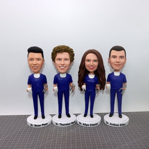 May include: Four custom-made figurines of people wearing blue scrubs and holding a wrench and a tooth. The figurines are standing on white bases with names printed on them: Dr. Dean Phan, Dr. Thurston Nash, Dr. Allyn Clause, and Dr. Joshua Allen.