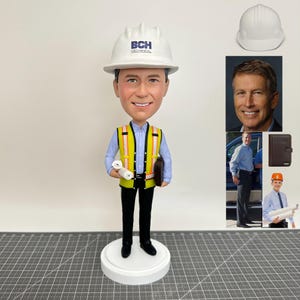 以下が含まれることがあります： 「BCH MECHANICAL」とプリントされた白いヘルメットをかぶった、建設作業員の服装をした男性のボブルヘッドフィギュア。丸められた青写真と茶色のポートフォリオを持っています。フィギュアは白い台座の上にあります。