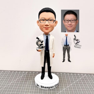 Könnte beinhalten: Eine personalisierte Bobblehead-Figur einer Person, die einen weißen Laborkittel trägt und ein Mikroskop hält. Die Figur steht auf einem weißen Sockel mit dem Namen "Dr. Shanyan Cho" darauf.