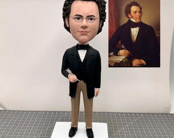 May include: A custom bobblehead figurine of a man with brown hair and glasses wearing a black suit and brown pants. The figurine is holding a quill pen in his right hand. A portrait of the man is shown in the background.
