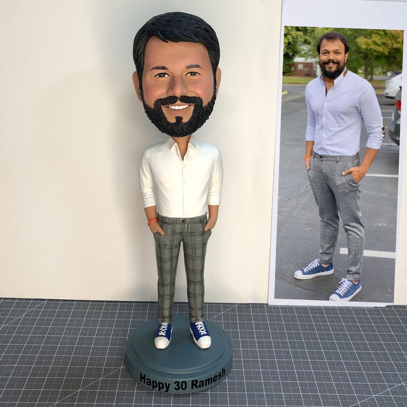 Może przedstawiać: Spersonalizowana figurka bobblehead przedstawiająca osobę z brodą, ubraną w białą koszulę i szare spodnie w kratę. Figurka stoi na okrągłej podstawie z napisem "Happy 30 Ramesh".