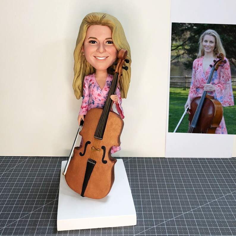 May include: A custom-made figurine of a person holding a cello. The figurine is wearing a pink and white floral dress. The cello is brown with a black bow.