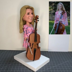 May include: A custom-made figurine of a person holding a cello. The figurine is wearing a pink dress with a floral pattern. The cello is brown with a black bow. The figurine is standing on a white base.