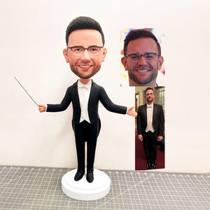 Könnte beinhalten: Eine maßgefertigte Figur eines Dirigenten in einem schwarzen Smoking, weißem Hemd und Fliege, der einen Taktstock hält. Die Figur hat einen karikaturartigen Kopf mit Brille und einem lächelnden Gesichtsausdruck. Inklusive Foto.