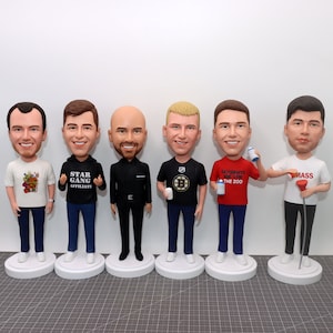 May include: Six custom bobbleheads of men wearing different shirts and holding various items. The bobbleheads are standing on a brown surface.