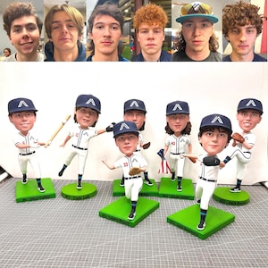 May include: A collection of baseball bobbleheads featuring young men in white and navy uniforms, each with a baseball cap. The bobbleheads stand on green bases, with names and numbers visible. Above, a row of the young men's faces.