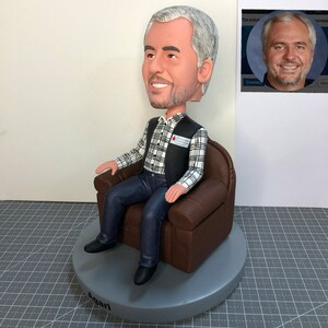 May include: A custom bobblehead figurine of a person sitting in a brown leather armchair. The person is wearing a black vest over a white and black plaid shirt and blue jeans. The bobblehead has a gray base with the name "Angari" written on it. The bobblehead has a red and white sticker on the vest that says "BLOOD WORKS FOUNDATION".