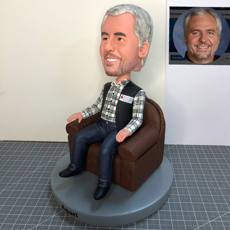 May include: A custom bobblehead figurine of a person sitting in a brown leather armchair. The person is wearing a black vest over a white and black plaid shirt and blue jeans. The bobblehead has a gray base with the name "Angari" written on it. The bobblehead has a red and white sticker on the vest that says "BLOOD WORKS FOUNDATION".
