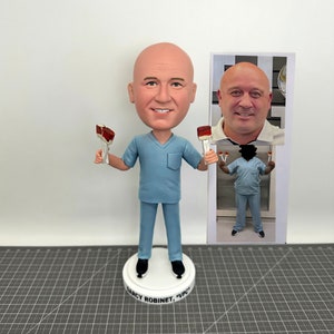 May include: A custom bobblehead figurine of a person wearing a light blue medical scrubs with a nameplate that reads "Darcy Robinett, VPCH". The figurine is holding two red and white paintbrushes.