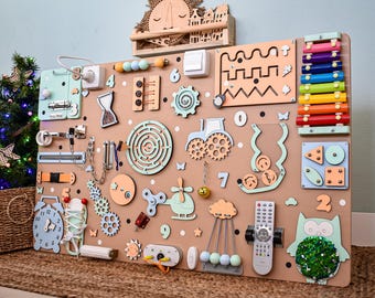 Puede incluir: Un gran tablero de actividades de madera con varios elementos interactivos, incluyendo un xilófono colorido, un reloj, un laberinto, engranajes, un mando a distancia, un búho con lentejuelas, un helicóptero y números pintados en tonos pastel.