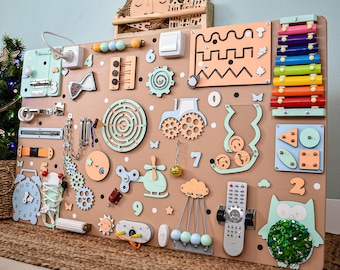 Könnte beinhalten: Ein großes, hölzernes Activity-Board mit verschiedenen interaktiven Elementen für Kinder. Es enthält einen Lichtschalter, Zahnräder, ein Labyrinth, ein Xylophon und eine Uhr. Das Board ist mit pastellfarbenen Formen und Zahlen verziert, und oben befinden sich eine Holzsonne und eine Stadt.