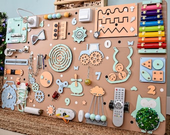 Könnte beinhalten: Eine große, hölzerne Beschäftigungstafel für Kleinkinder mit verschiedenen interaktiven Elementen. Enthält eine Uhr, einen Lichtschalter, Zahnräder, ein Labyrinth, ein Xylophon und andere sensorische Aktivitäten. Die Tafel ist hauptsächlich beige mit pastellfarbenen Akzenten.