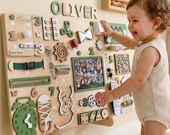 Puede incluir: Un tablero sensorial de madera con varios elementos interactivos, incluyendo engranajes, pestillos, una cremallera y un reloj. El tablero presenta el nombre "OLIVER" en letras verdes y una pequeña imagen enmarcada. Diseñado para el desarrollo infantil temprano.
