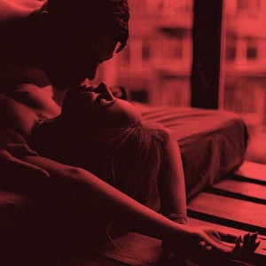 May include: A romantic scene bathed in red light. A person leans over another, their faces close. The image evokes intimacy and closeness, with a focus on the figures' interaction. The setting appears to be a bedroom.