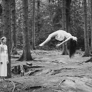 May include: Monochrome image of a forest with two figures. One stands by a tree, the other floats in the air. Both are dressed in white. The ground is covered in leaves and tree roots. The scene evokes a sense of mystery.