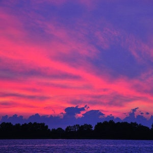 May include: A vibrant sunset over a body of water. The sky is filled with streaks of pink and red clouds against a backdrop of deep blue. A dark silhouette of trees and land is visible at the horizon.