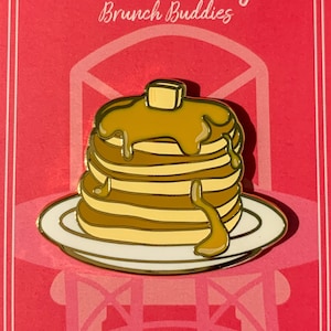 May include: A gold-colored enamel pin featuring a stack of pancakes with syrup and a pat of butter on top. The pin is on a pink background with the text "Hobnob Lifestyle Brunch Buddies" and the social media handle "@HobnobLifestyle".