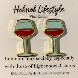 May include: A pair of gold-tone earrings shaped like wine glasses. The glasses are filled with red wine and have a light blue background. The earrings are on a white card with the text "Hobnob Lifestyle Vino Edition" and "hob.nob | mix socially, especially with those of higher social status @HobNobLifestyle | HobNobLifestyle.com".