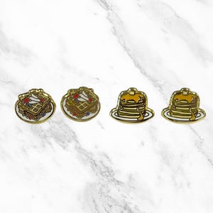 May include: Four enamel pins featuring breakfast foods. Two pins are of waffles with whipped cream and berries, and two pins are of pancakes with syrup.