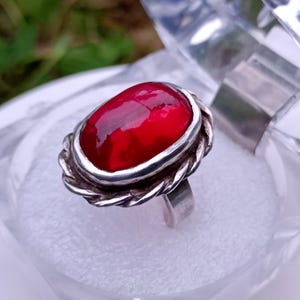 May include: A silver ring with a large, oval, red gemstone. The gemstone is set in a silver bezel with a twisted rope design around it. The ring is displayed in a clear plastic box.