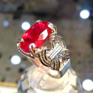 May include: A silver ring with a large, oval-shaped, red gemstone set in the center. The ring is made of wire that is twisted and woven together.